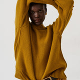 A model wearing a mustard yellow ribbed knit sweater and brown trousers with a black belt, posing against a plain light gray background.