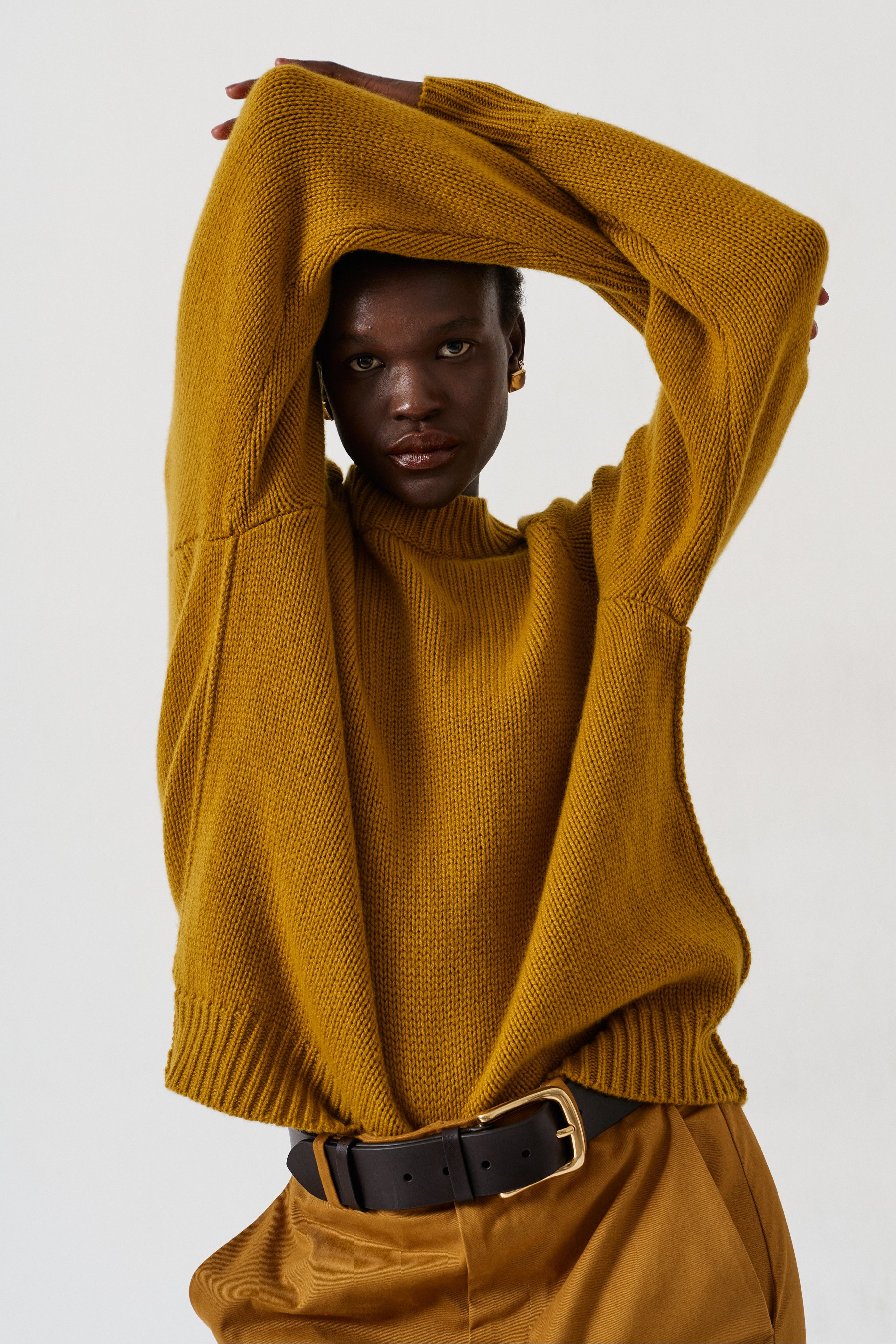 A model wearing a mustard yellow ribbed knit sweater and brown trousers with a black belt, posing against a plain light gray background.