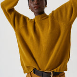 A model wearing a mustard yellow ribbed knit mock-neck sweater and matching trousers, accessorized with a black belt, against a plain light gray background.