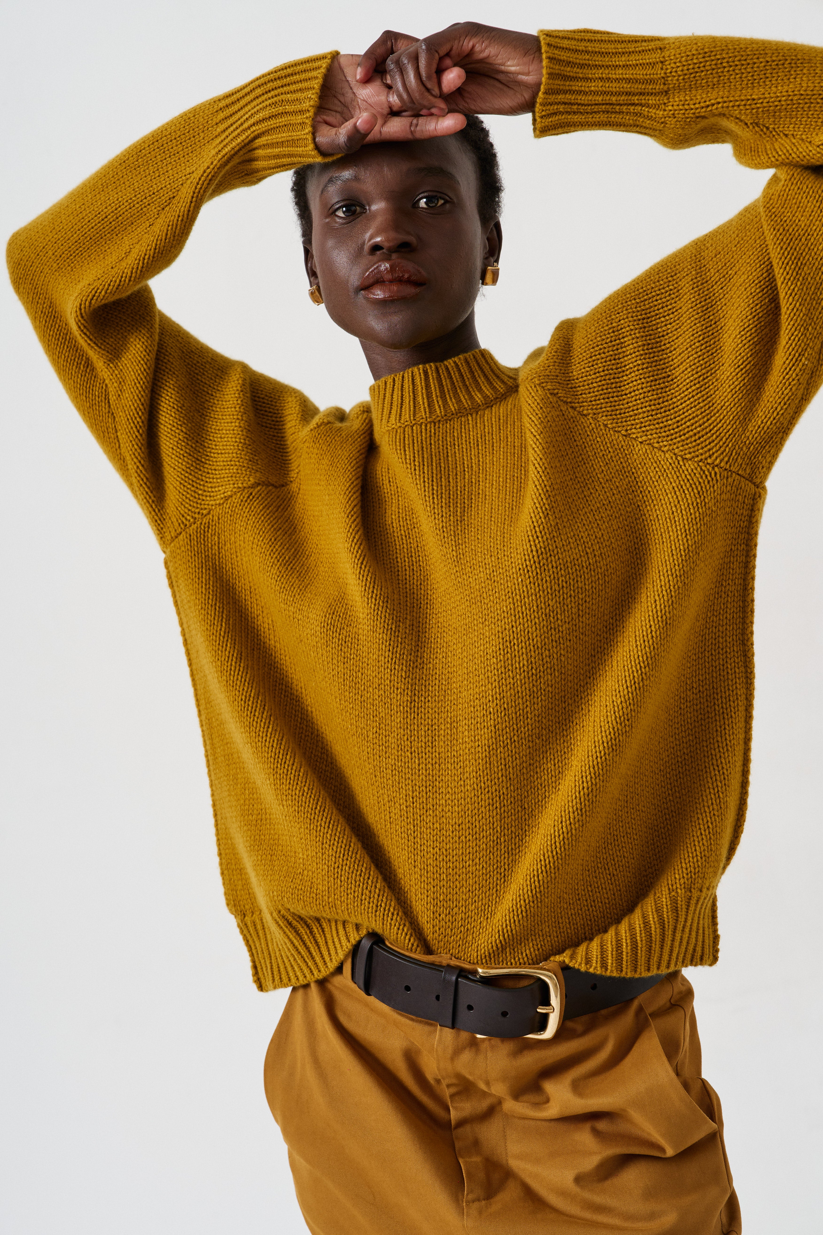 A model wearing a mustard yellow ribbed knit mock-neck sweater and matching trousers, accessorized with a black belt, against a plain light gray background.