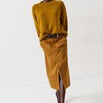 A model walks in a studio wearing a mustard yellow ribbed knit sweater and a matching mustard yellow midi skirt with a front slit, styled with a black belt and dark brown pointed-toe boots.