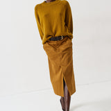 A model walks in a studio wearing a mustard yellow ribbed knit sweater and a matching mustard yellow midi skirt with a front slit, styled with a black belt and dark brown pointed-toe boots.