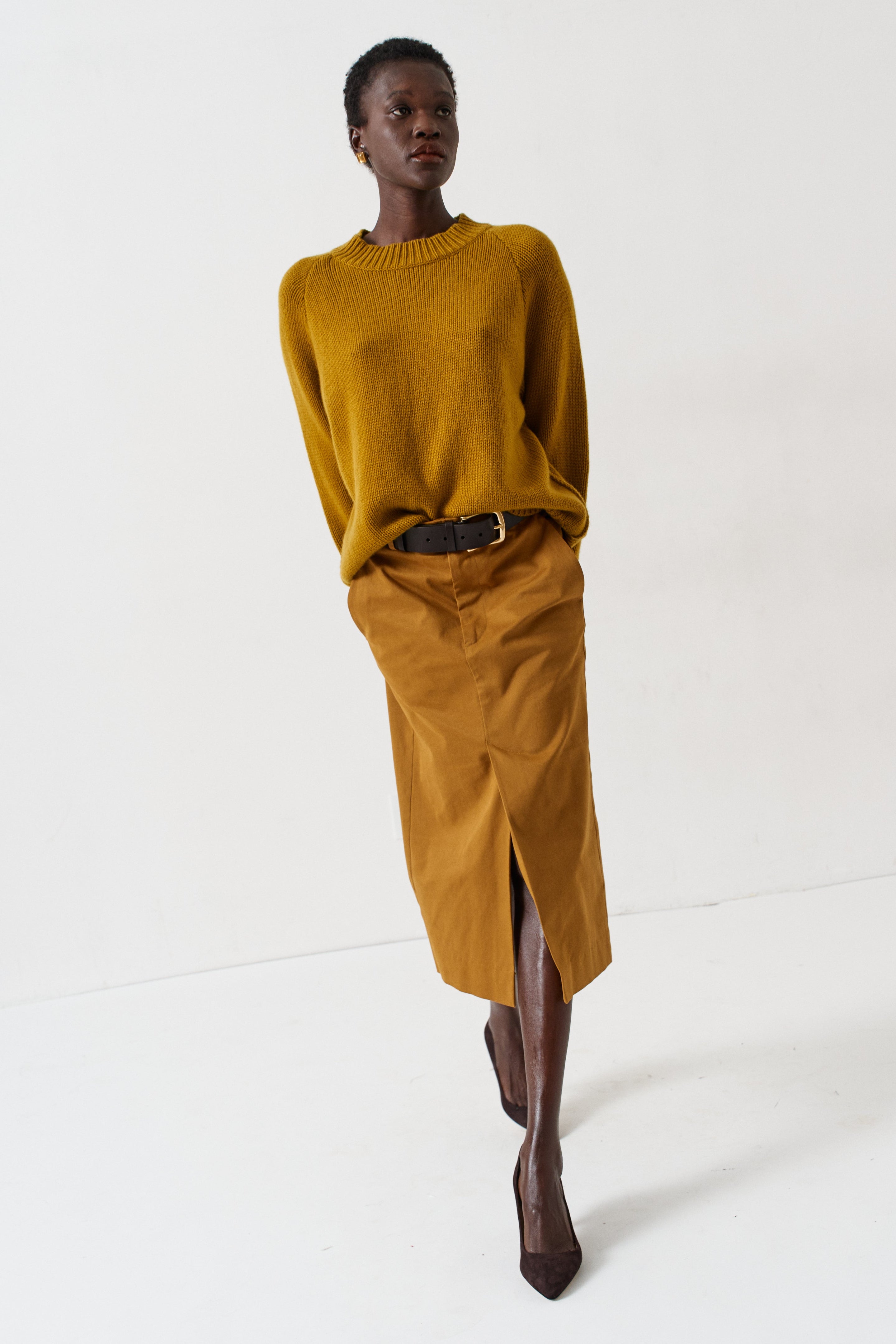 A model walks in a studio wearing a mustard yellow ribbed knit sweater and a matching mustard yellow midi skirt with a front slit, styled with a black belt and dark brown pointed-toe boots.