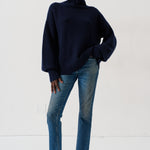 A model wearing a dark navy blue turtleneck sweater, blue jeans, and dark loafers, standing against a plain white studio background.