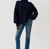 A model wearing a dark navy blue turtleneck sweater, blue jeans, and dark loafers, standing against a plain white studio background.