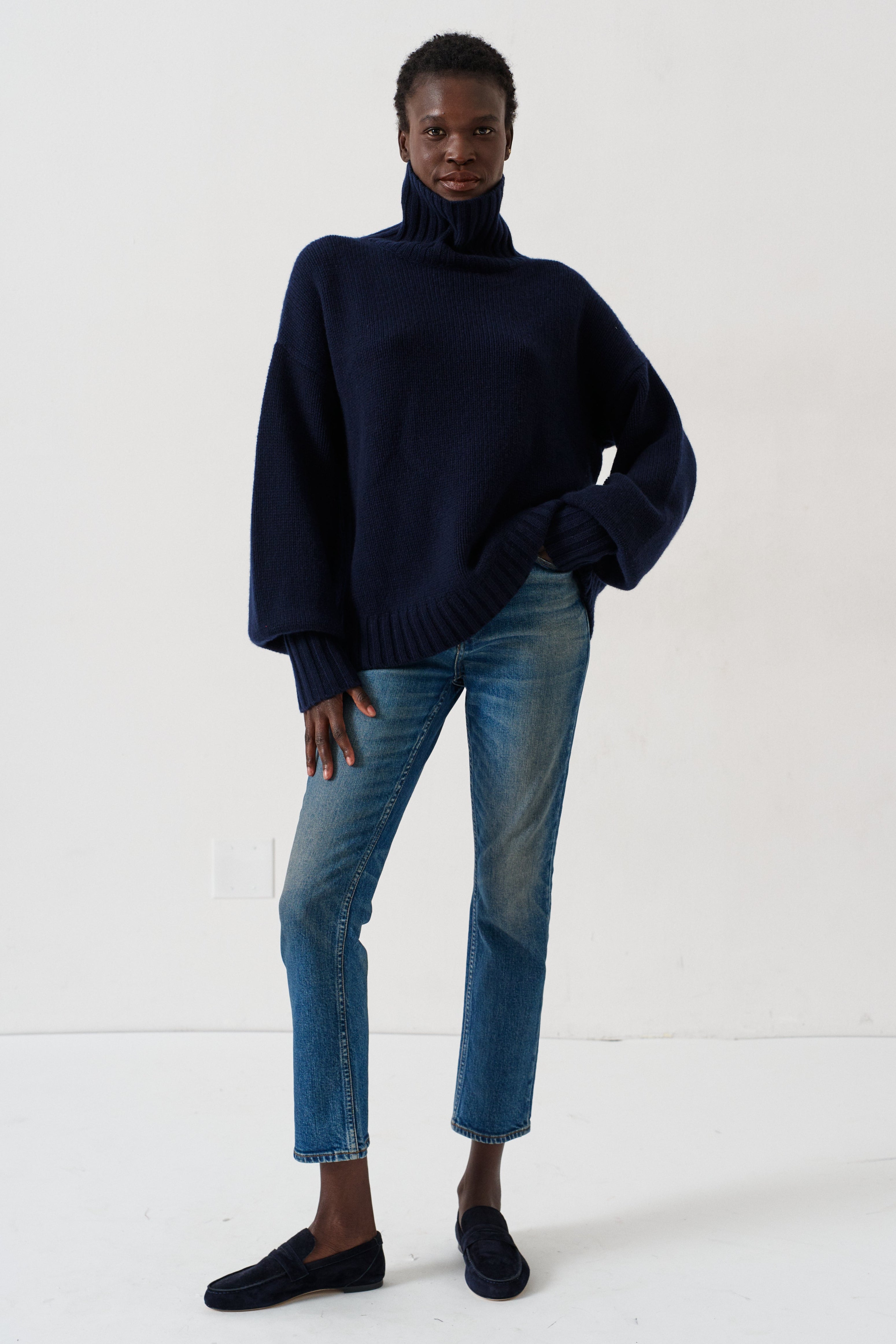A model wearing a dark navy blue turtleneck sweater, blue jeans, and dark loafers, standing against a plain white studio background.