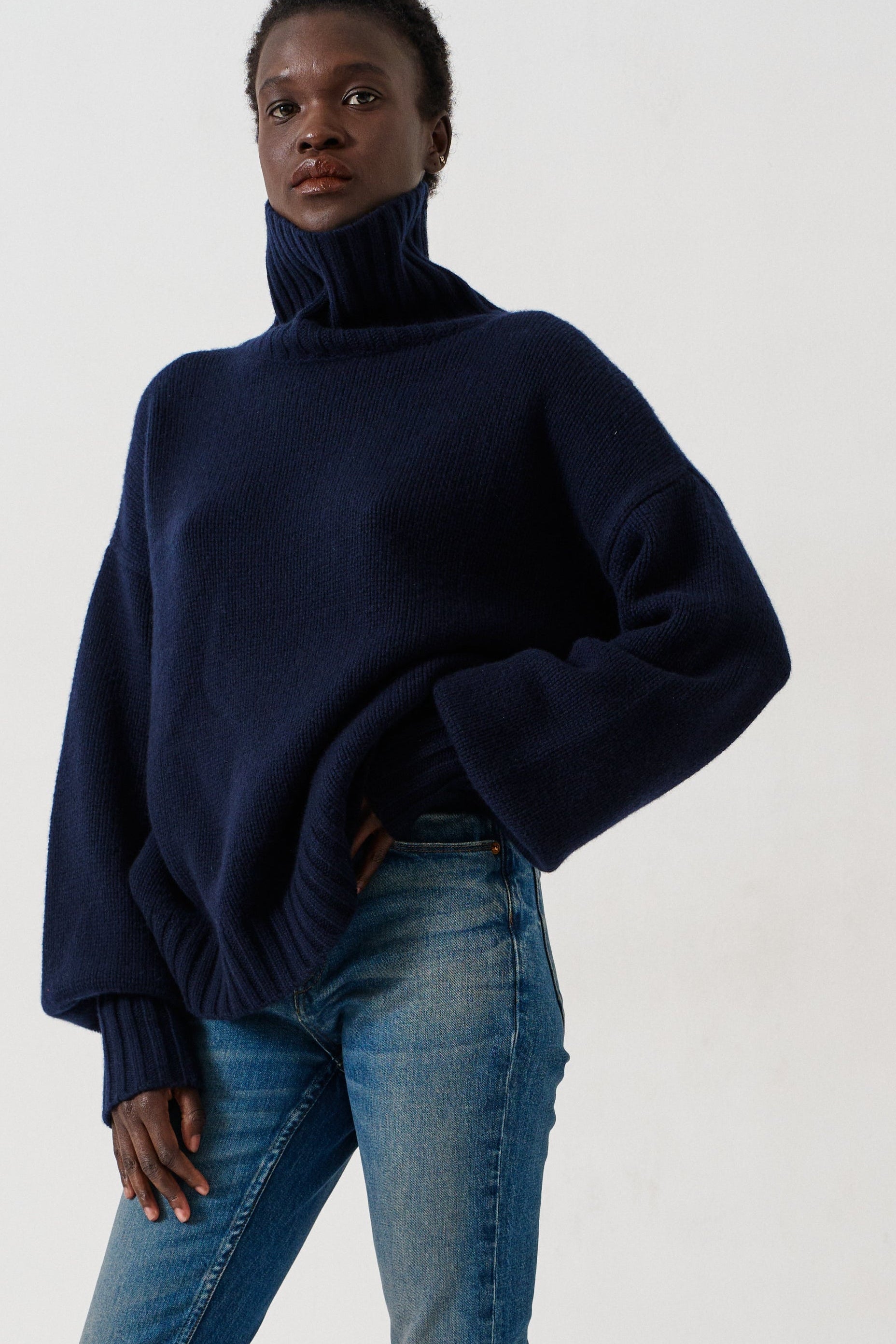 A model wearing a dark navy blue turtleneck sweater, blue cropped jeans, and dark slip-on shoes, standing against a plain white studio background.
