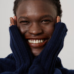 A close-up portrait of a smiling person with short dark hair, wearing a dark navy blue ribbed knit sweater with their hands tucked into the collar, against a plain light gray background.