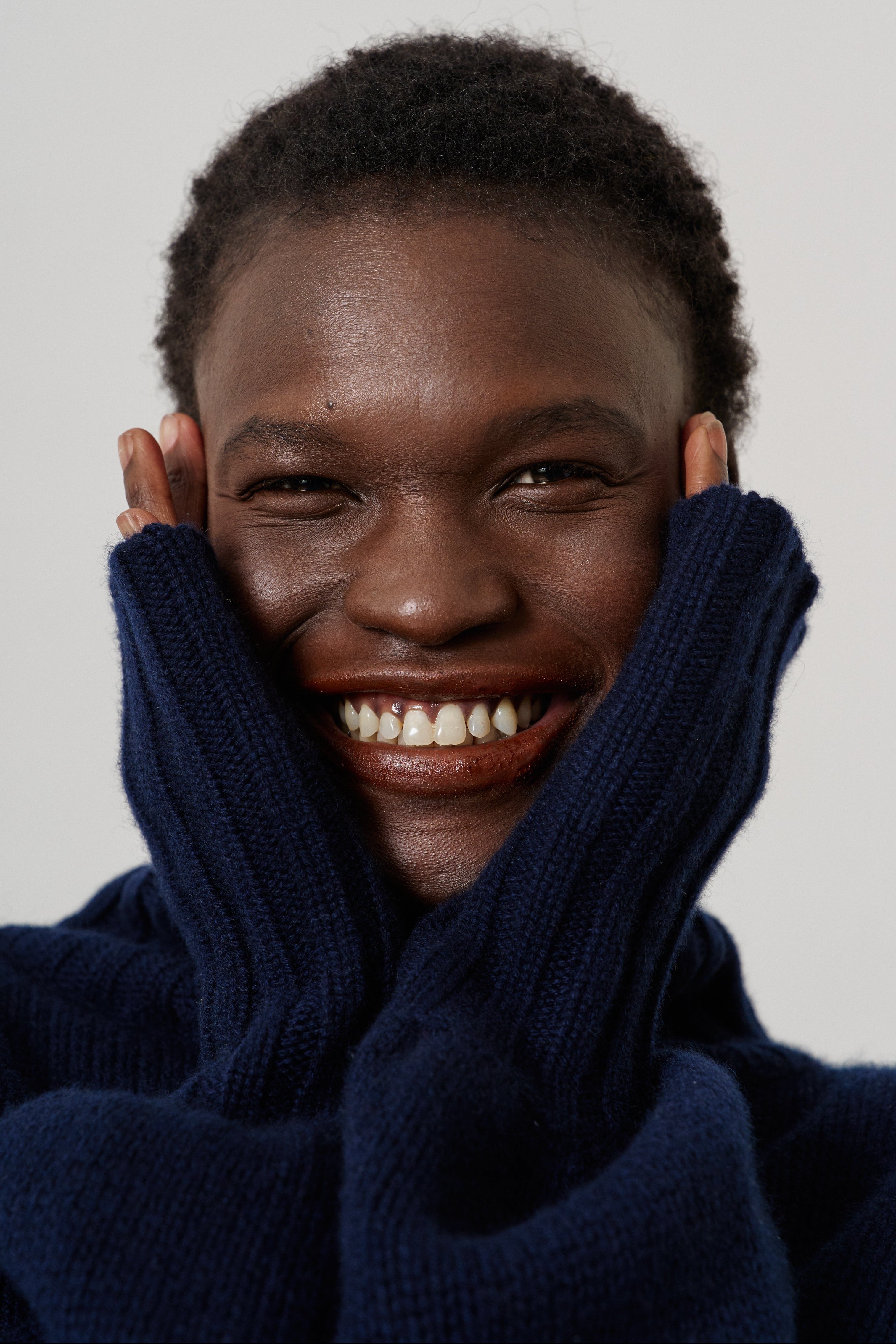 A close-up portrait of a smiling person with short dark hair, wearing a dark navy blue ribbed knit sweater with their hands tucked into the collar, against a plain light gray background.