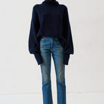 A model stands against a plain white studio background wearing a dark navy blue turtleneck sweater, blue jeans, and dark loafers.