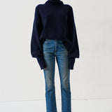 A model stands against a plain white studio background wearing a dark navy blue turtleneck sweater, blue jeans, and dark loafers.