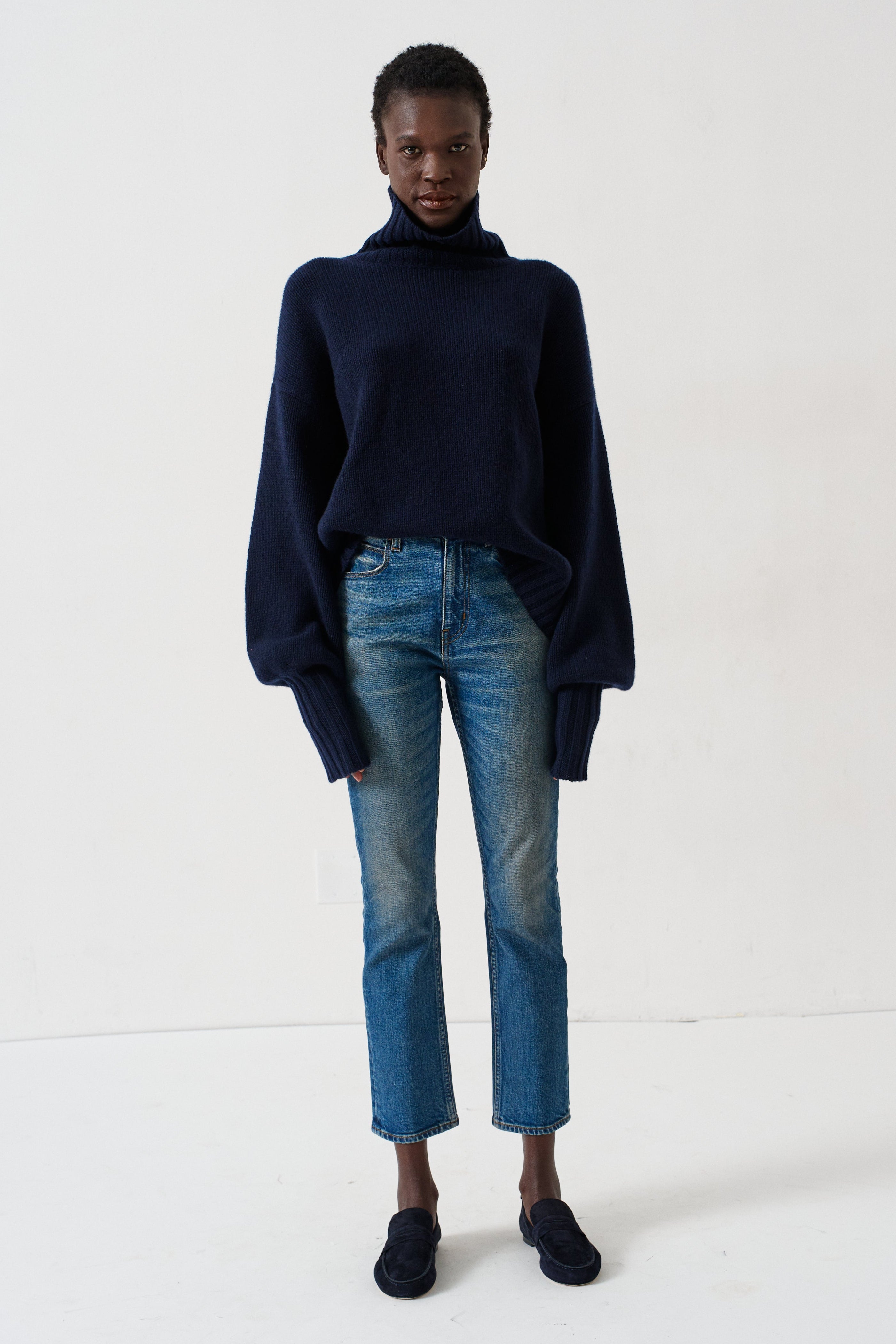 A model stands against a plain white studio background wearing a dark navy blue turtleneck sweater, blue jeans, and dark loafers.