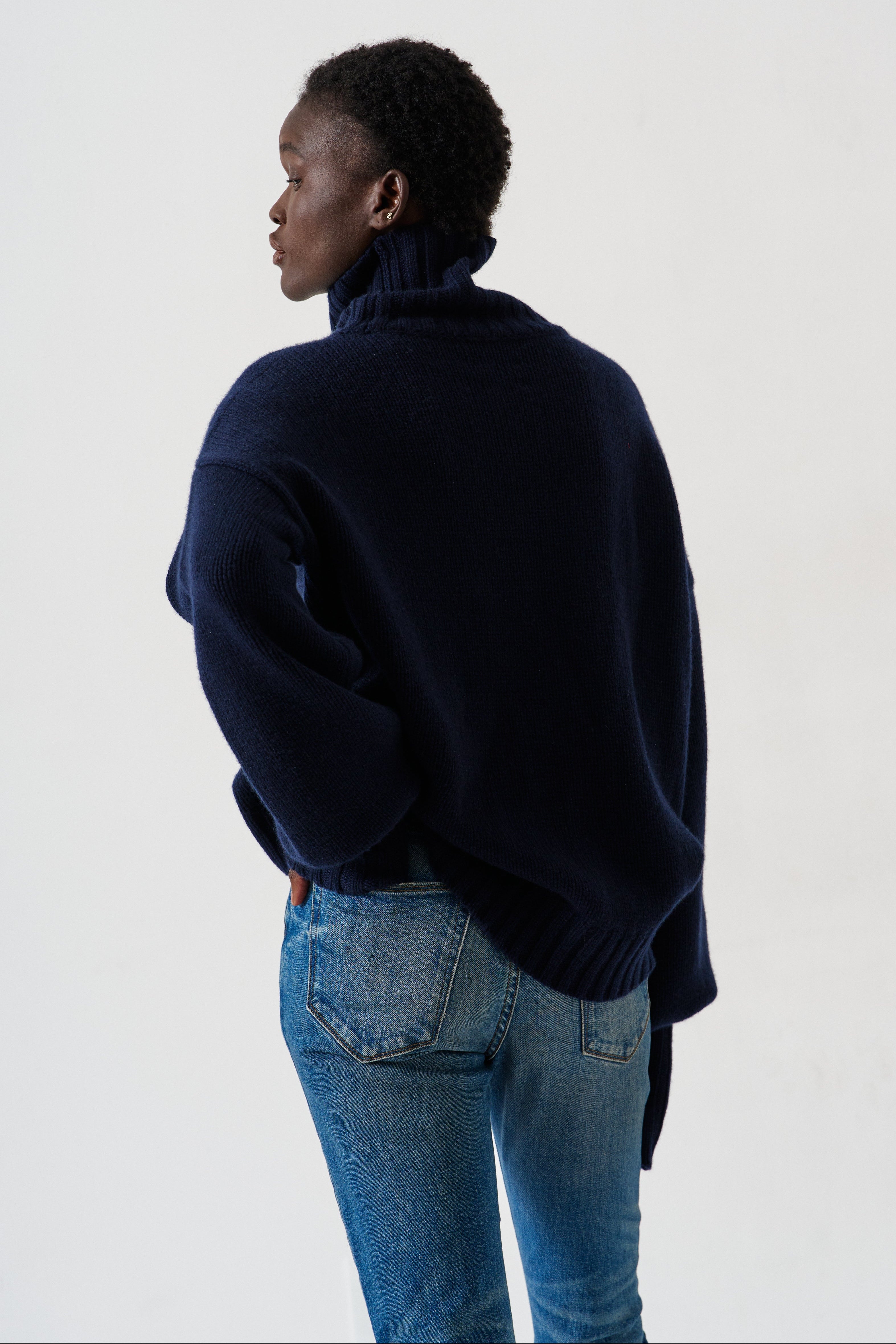 A person shown from the back wearing an oversized navy blue turtleneck sweater and blue jeans against a plain light gray studio background.