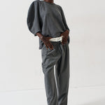 A model stands in a studio wearing a dark gray two-piece set consisting of a loose, voluminous long-sleeve top and matching wide-leg trousers, styled with a white belt and bright pink pointed-toe heels.