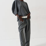 A model stands in a studio wearing a dark gray two-piece set consisting of a loose, voluminous long-sleeve top and matching wide-leg trousers, styled with a white belt and bright pink pointed-toe heels.