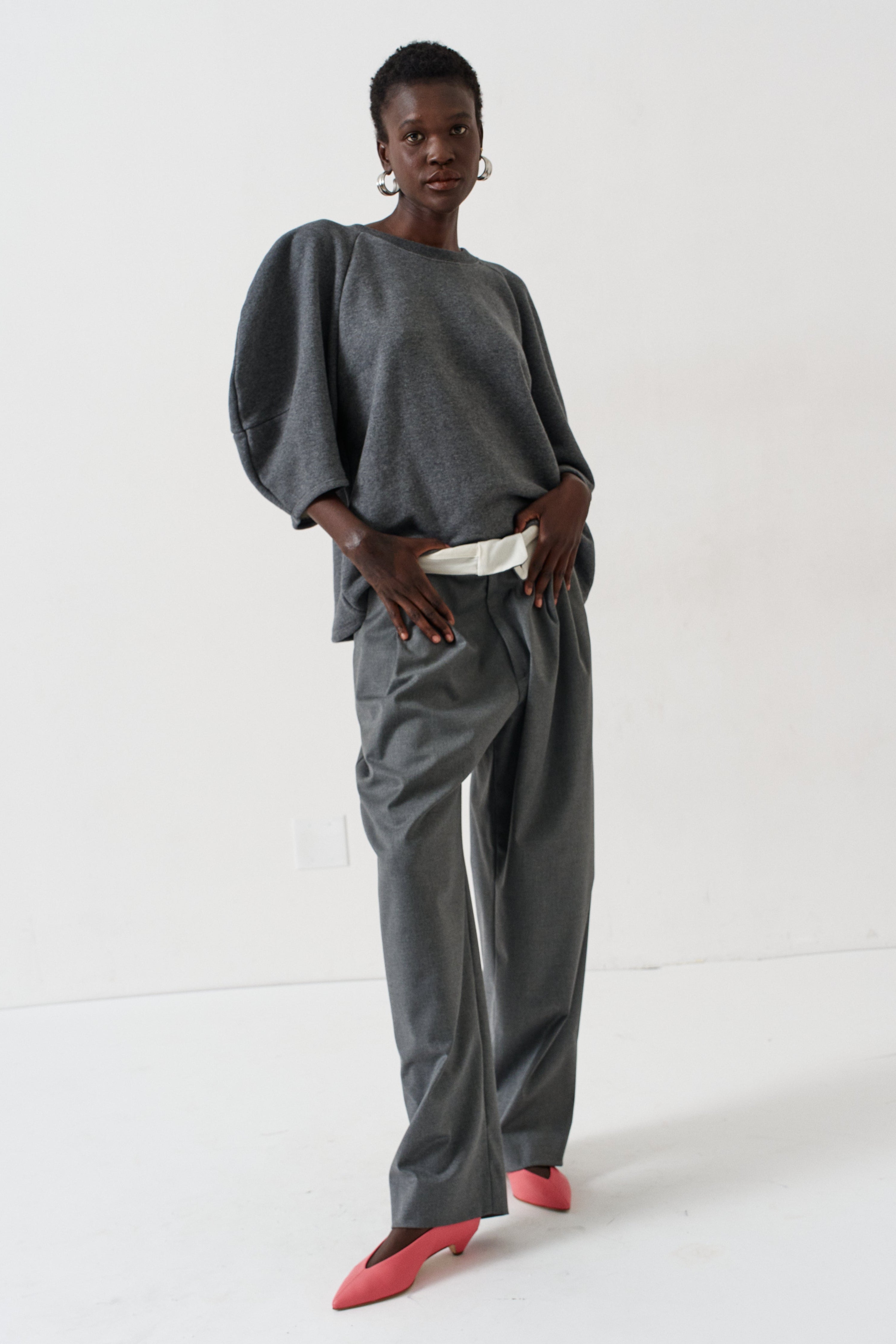 A model stands in a studio wearing a dark gray two-piece set consisting of a loose, voluminous long-sleeve top and matching wide-leg trousers, styled with a white belt and bright pink pointed-toe heels.