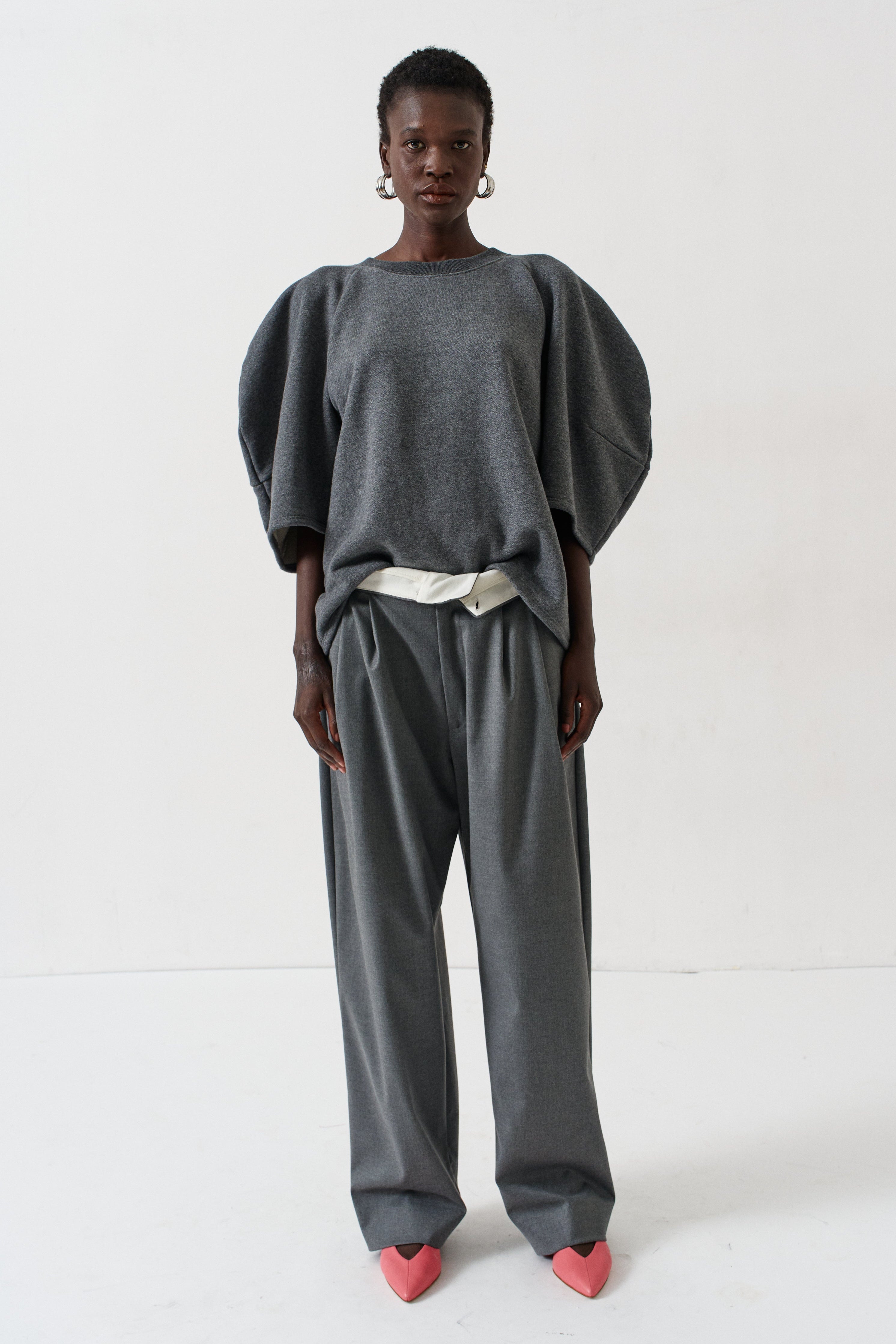 A model stands against a plain white studio background wearing a dark gray two-piece outfit consisting of a short puff-sleeve top and matching wide-leg trousers, styled with a cream ribbon at the waist and pink heart-shaped shoes.