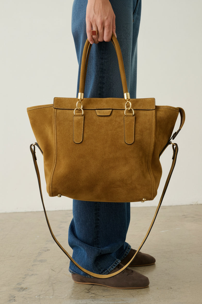 A person wearing blue jeans and brown shoes holds a large tan suede tote bag with gold-tone hardware and a long shoulder strap in a minimalist studio setting.