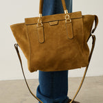 A person wearing blue jeans and brown shoes holds a large tan suede tote bag with gold-tone hardware and a long shoulder strap in a minimalist studio setting.