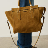 A person wearing blue jeans and brown shoes holds a large tan suede tote bag with gold-tone hardware and a long shoulder strap in a minimalist studio setting.
