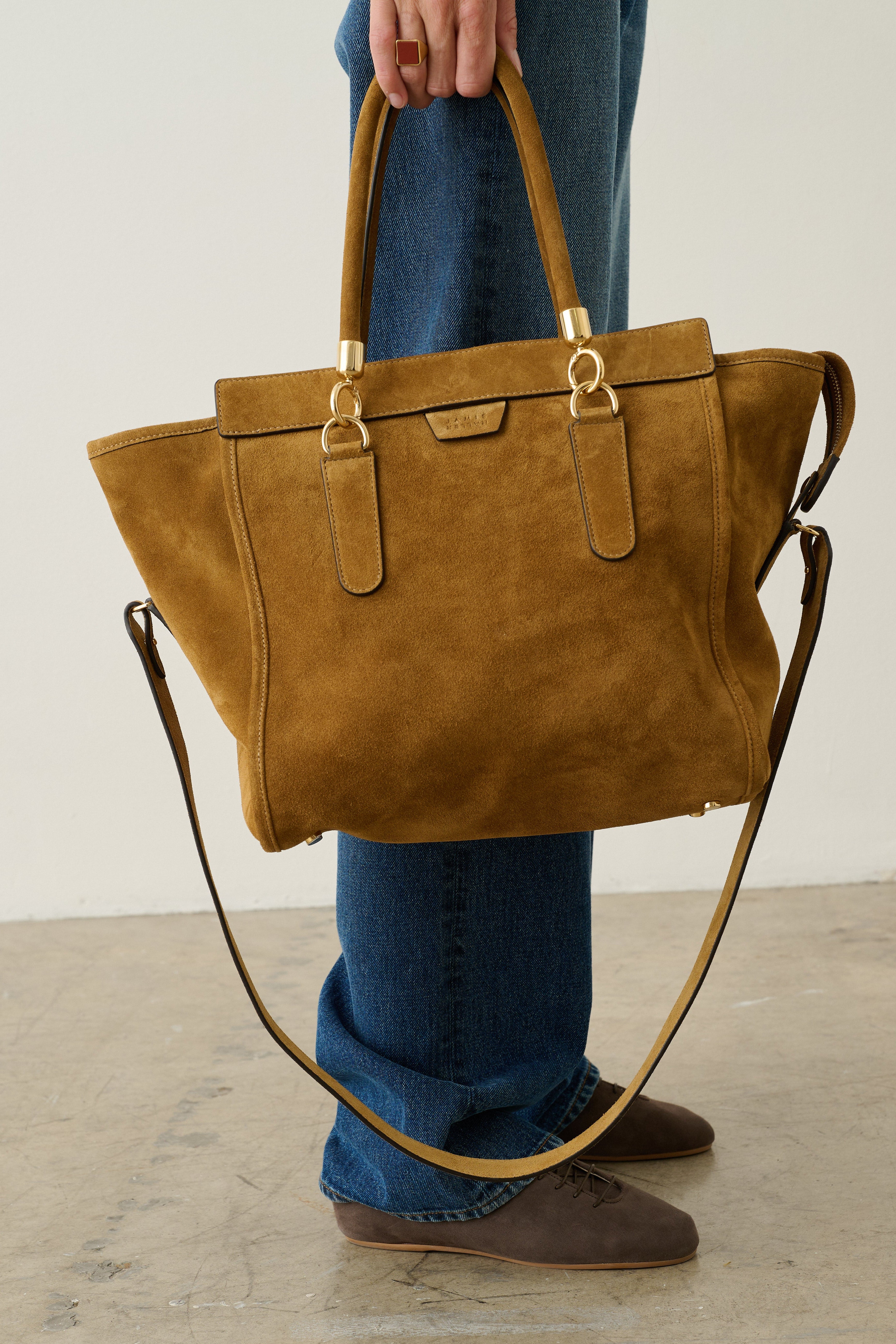 A person wearing blue jeans and brown shoes holds a large tan suede tote bag with gold-tone hardware and a long shoulder strap in a minimalist studio setting.