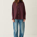 A woman stands against a plain off-white studio background wearing a long-sleeve button-up shirt with vertical dark red and blue stripes, blue jeans, and brown shoes.