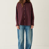 A woman stands against a plain off-white studio background wearing a long-sleeve button-up shirt with vertical dark red and blue stripes, blue jeans, and brown shoes.