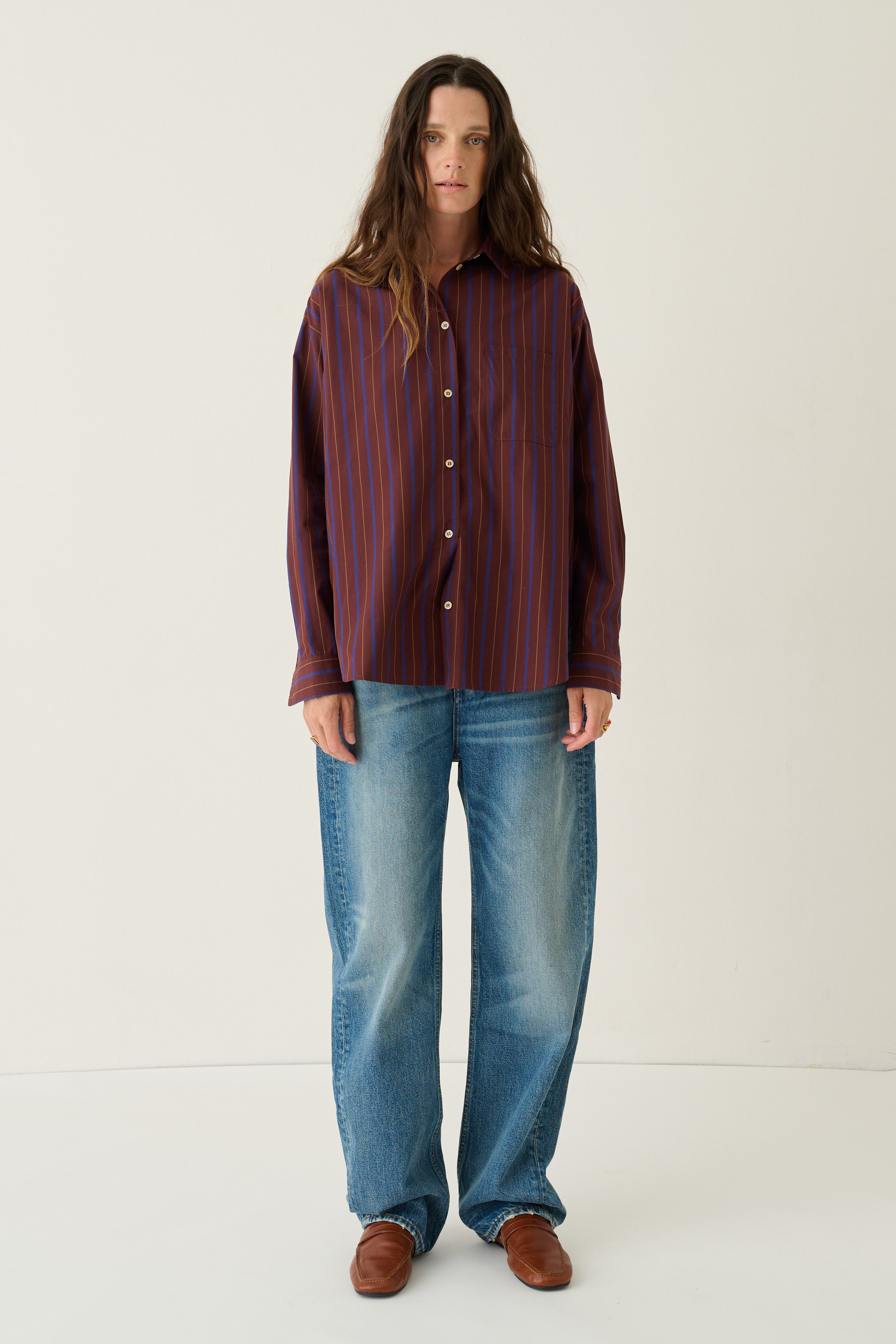 A woman stands against a plain off-white studio background wearing a long-sleeve button-up shirt with vertical dark red and blue stripes, blue jeans, and brown shoes.