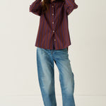 A woman stands in a white studio setting wearing a dark red and navy vertically striped button-up shirt, blue jeans, and brown shoes.