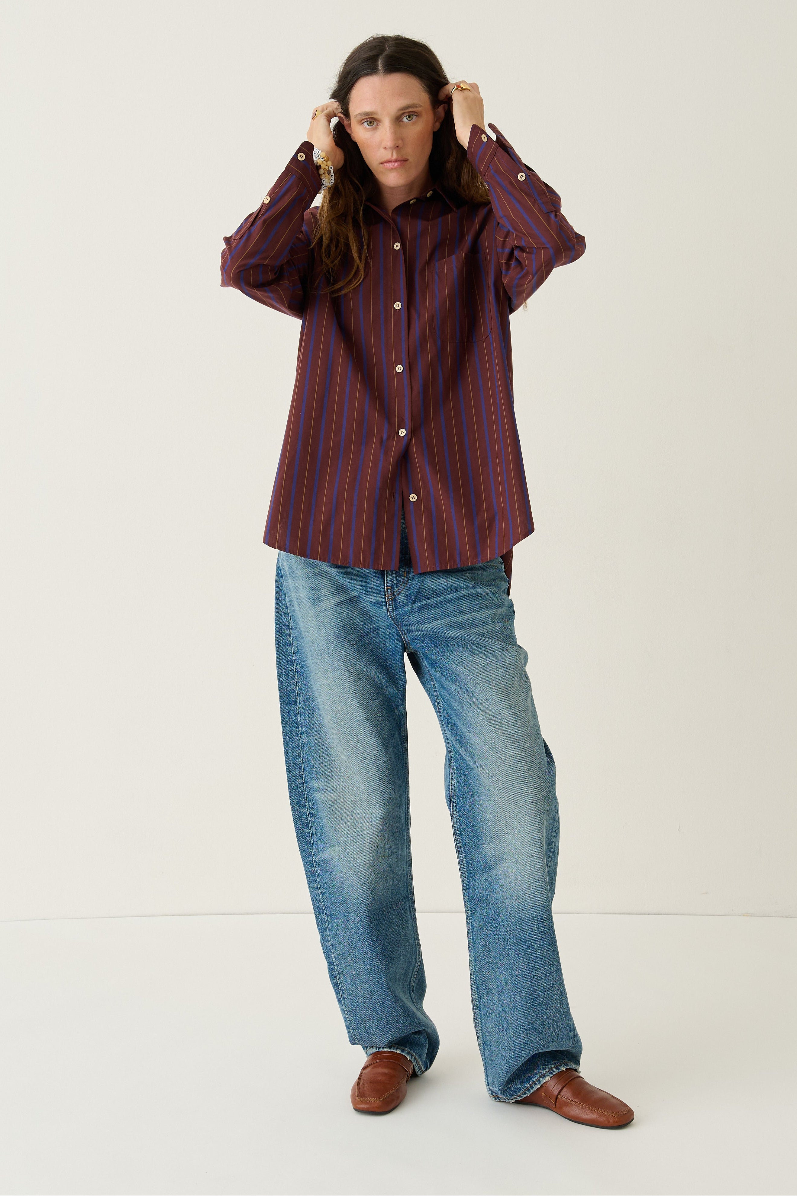 A woman stands in a white studio setting wearing a dark red and navy vertically striped button-up shirt, blue jeans, and brown shoes.