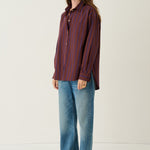A woman stands in a studio wearing a long-sleeve button-up shirt with vertical red and blue stripes, blue jeans, and brown shoes against a plain off-white background.