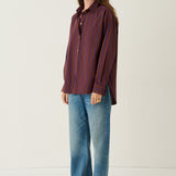 A woman stands in a studio wearing a long-sleeve button-up shirt with vertical red and blue stripes, blue jeans, and brown shoes against a plain off-white background.