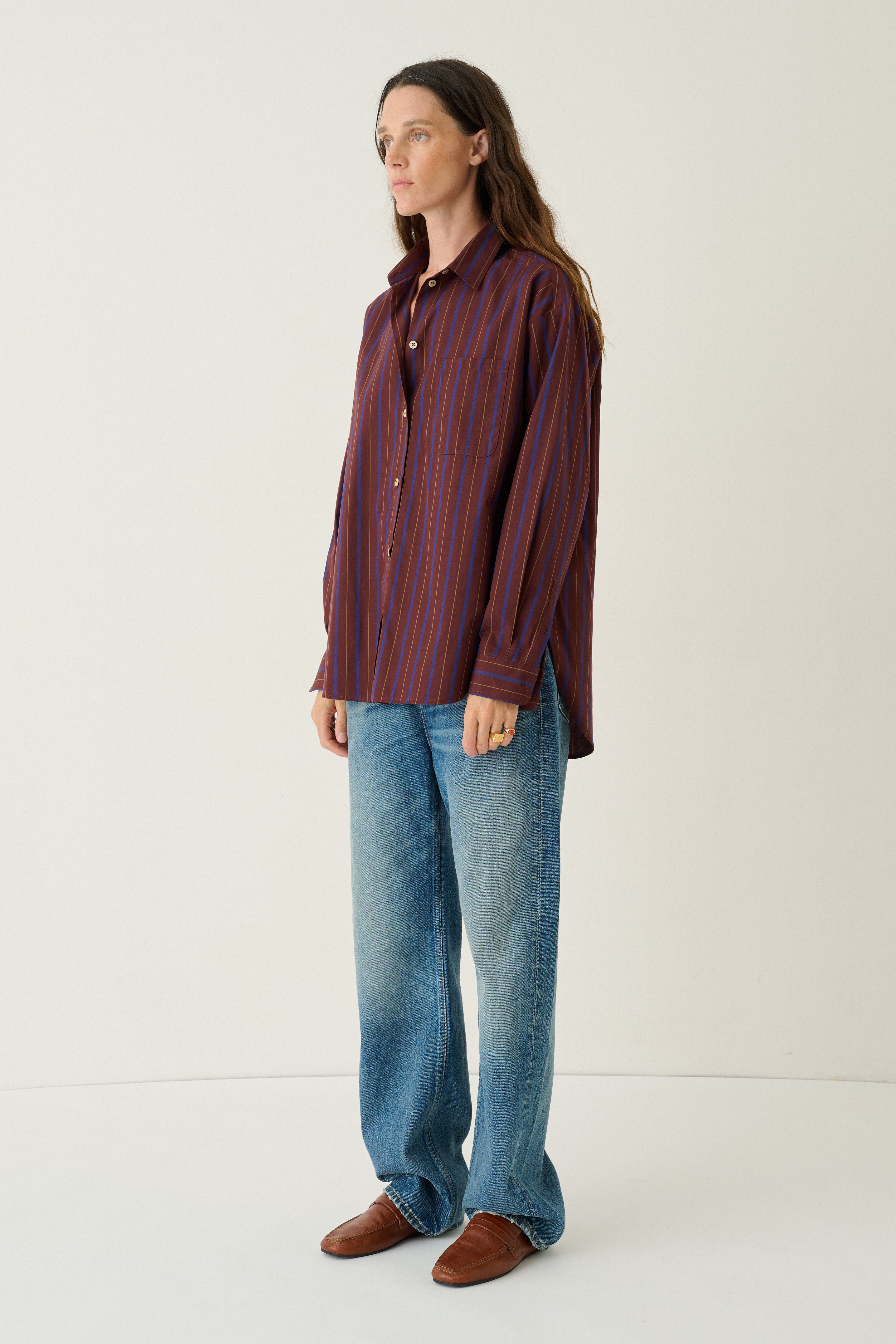A woman stands in a studio wearing a long-sleeve button-up shirt with vertical red and blue stripes, blue jeans, and brown shoes against a plain off-white background.
