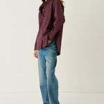 A woman modeling a long-sleeve button-up shirt with vertical burgundy and navy stripes, paired with light-wash blue jeans and brown leather shoes, standing against a plain off-white studio background.