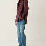 A woman modeling a long-sleeve button-up shirt with vertical burgundy and navy stripes, paired with light-wash blue jeans and brown leather shoes, standing against a plain off-white studio background.