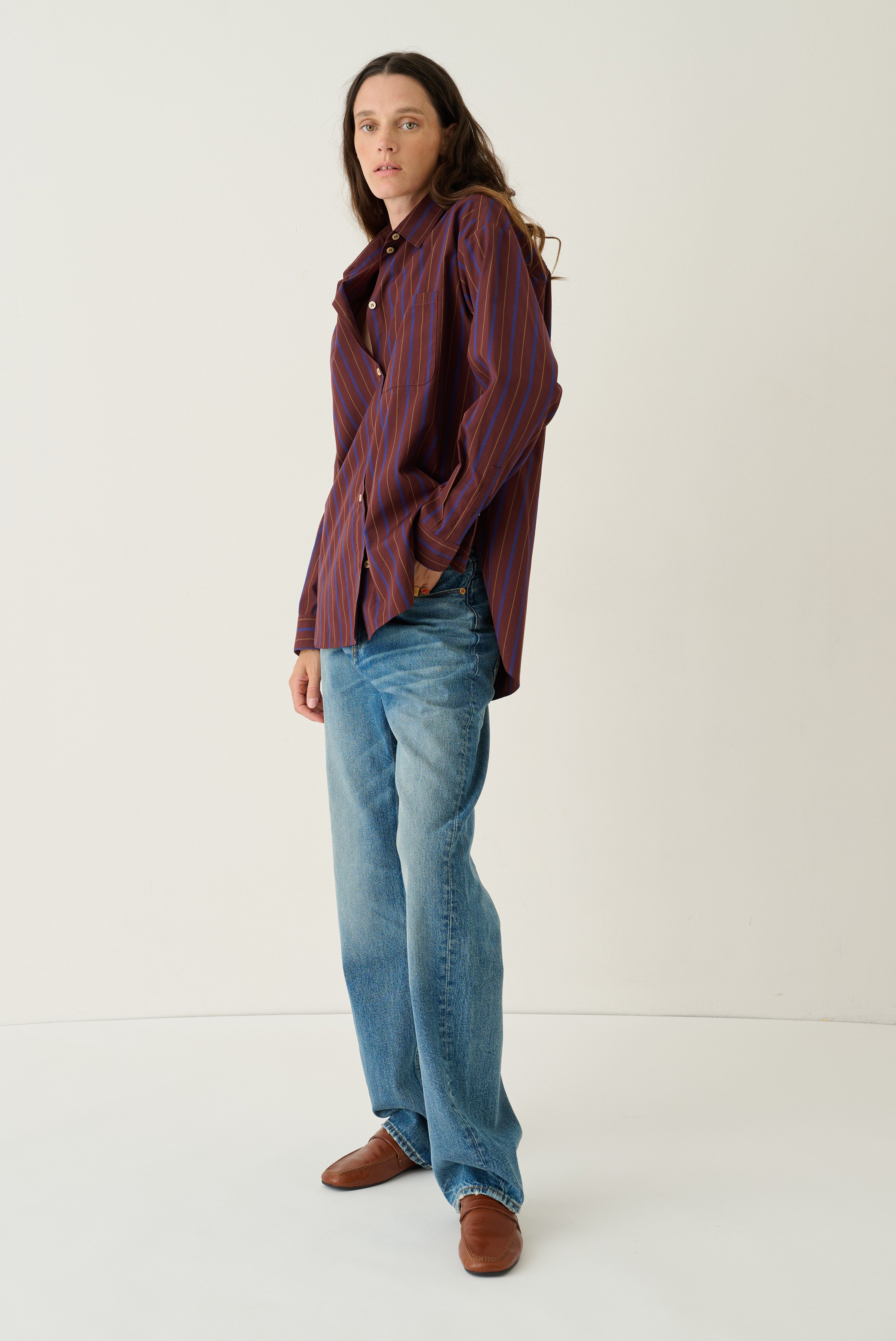 A woman modeling a long-sleeve button-up shirt with vertical burgundy and navy stripes, paired with light-wash blue jeans and brown leather shoes, standing against a plain off-white studio background.