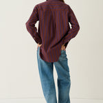 A person standing with their back to the camera, wearing a long-sleeve button-up shirt with vertical red and dark blue stripes, loose blue jeans, and brown shoes, against a plain off-white studio background.