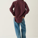 A person standing with their back to the camera, wearing a long-sleeve button-up shirt with vertical red and dark blue stripes, loose blue jeans, and brown shoes, against a plain off-white studio background.