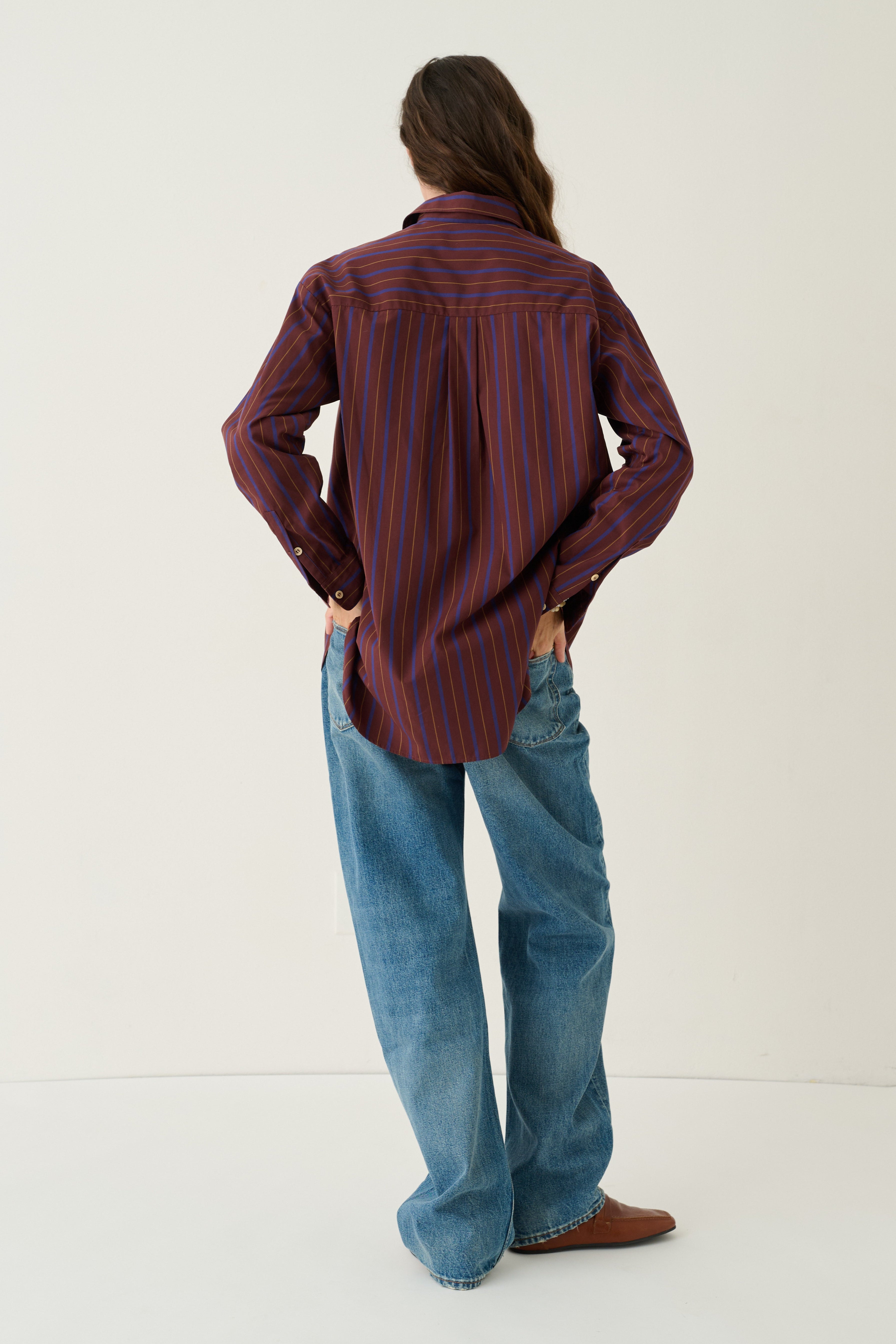 A person standing with their back to the camera, wearing a long-sleeve button-up shirt with vertical red and dark blue stripes, loose blue jeans, and brown shoes, against a plain off-white studio background.