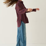 A woman in a side profile wearing a dark red and blue striped button-up shirt, wide-leg blue jeans, and brown leather shoes, standing against a plain off-white studio background.