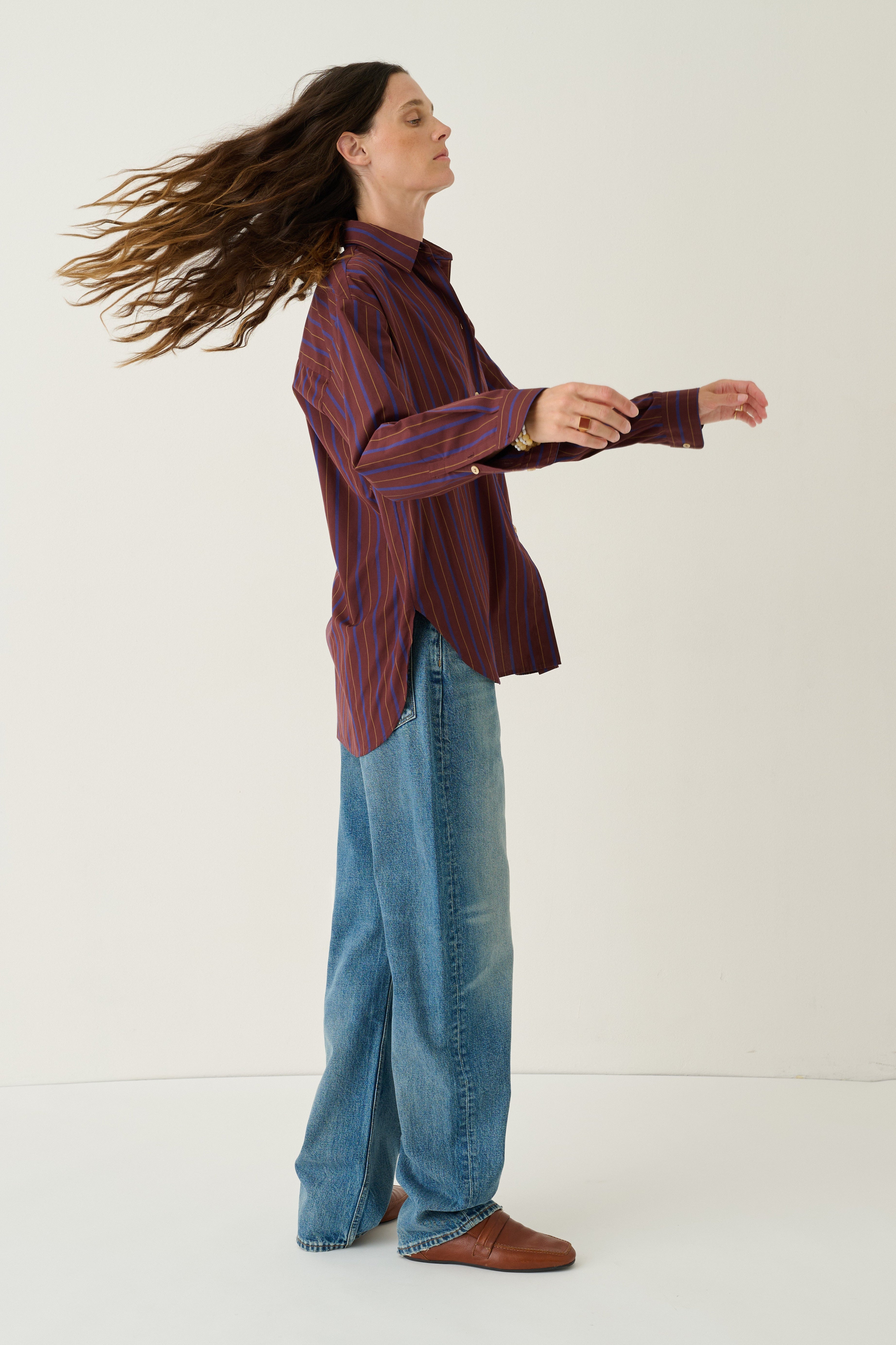 A woman in a side profile wearing a dark red and blue striped button-up shirt, wide-leg blue jeans, and brown leather shoes, standing against a plain off-white studio background.