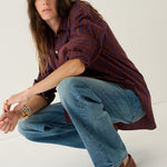 A woman crouching in a studio setting, wearing a dark red and navy vertically striped button-down shirt, wide-leg blue jeans, and brown leather loafers.