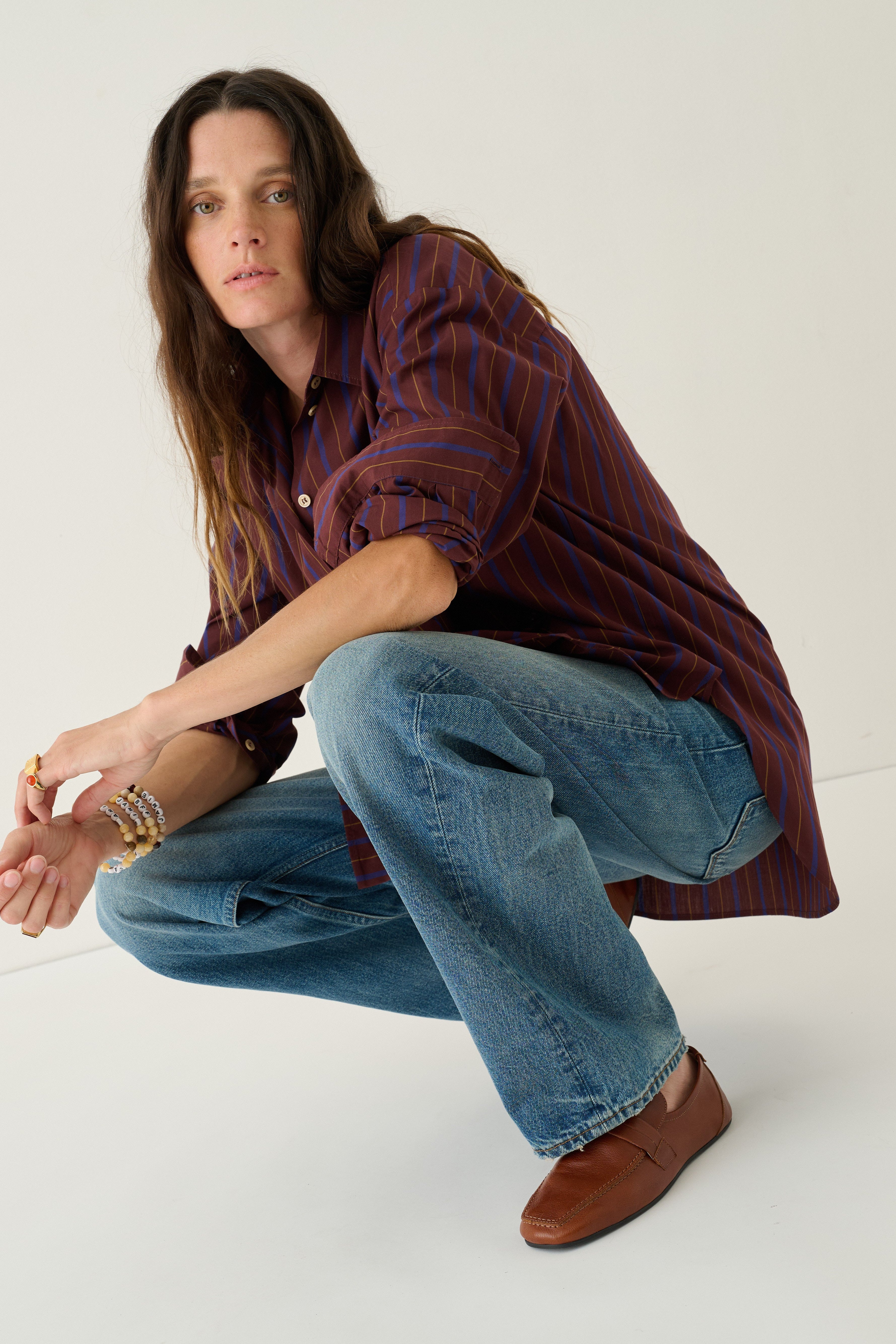 A woman crouching in a studio setting, wearing a dark red and navy vertically striped button-down shirt, wide-leg blue jeans, and brown leather loafers.