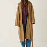 A woman stands against a plain off-white studio background wearing a long tan trench coat over a red and blue striped button-up shirt, blue jeans, and brown leather shoes.