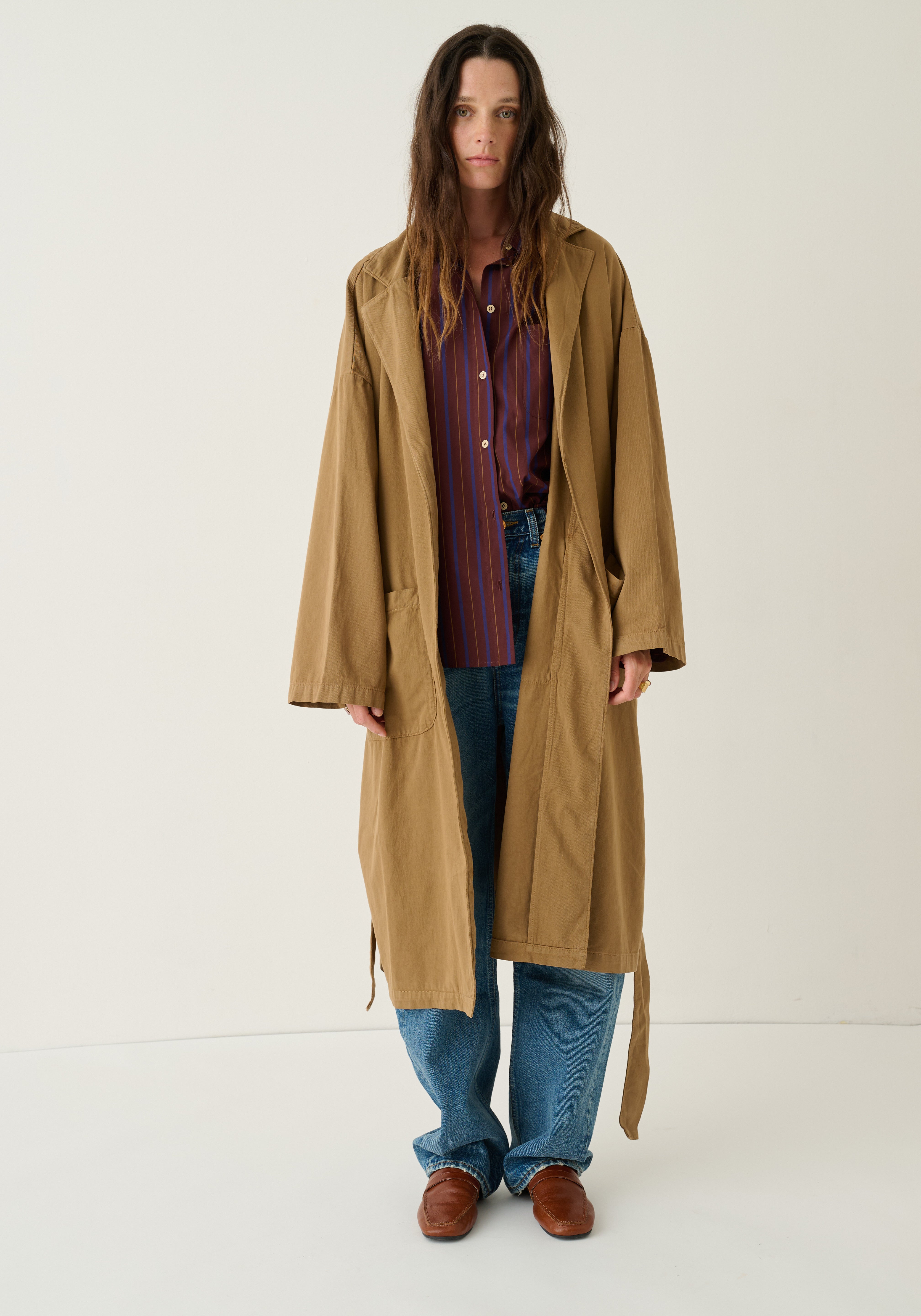 A woman stands against a plain off-white studio background wearing a long tan trench coat over a red and blue striped button-up shirt, blue jeans, and brown leather shoes.