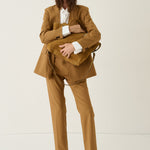 A model stands against a plain off-white studio background wearing a camel brown tailored suit with a white collared shirt, tan loafers, and holding a matching camel brown suede backpack.