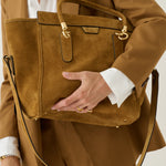 A person in a camel-brown blazer and matching trousers carries a large tan suede tote bag with gold-tone hardware and a black shoulder strap against a light neutral background.