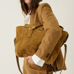 A model wearing a matching brown suit carries a large brown suede tote bag with gold-tone hardware and a white shirt underneath, set against a plain light background.