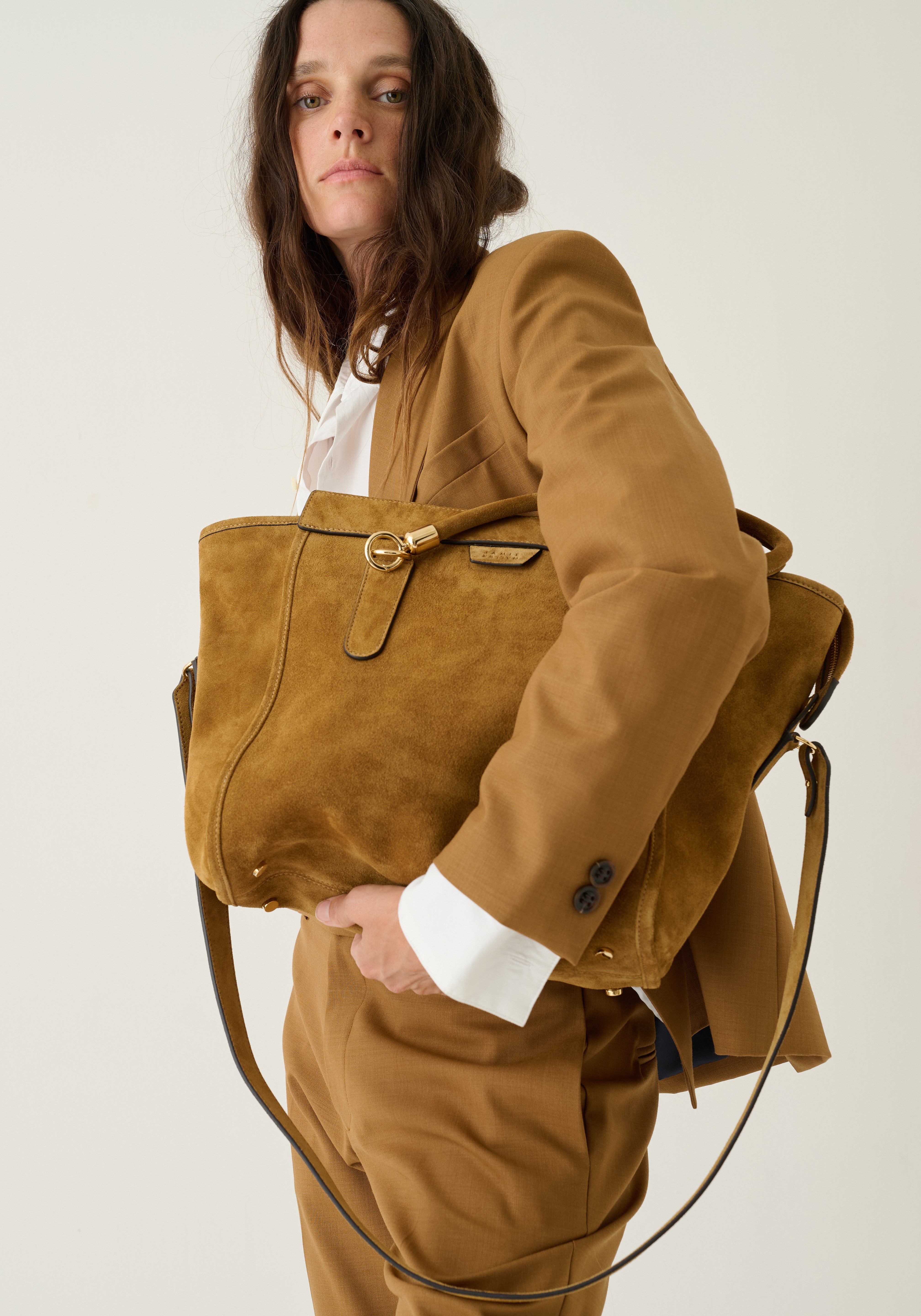A model wearing a matching brown suit carries a large brown suede tote bag with gold-tone hardware and a white shirt underneath, set against a plain light background.