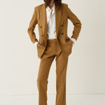 A model wearing a camel brown double-breasted blazer and matching trousers, styled with a white button-down shirt and tan suede shoes, standing against a plain off-white studio background.